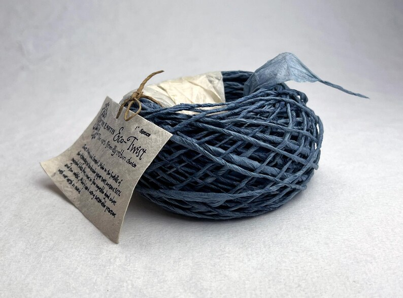 May include: A coil of blue paper twine labeled Eco-Twist in the color Spruce. The twine is wound into a circular shape and features a beige paper tag tied with a thin brown string. The tag includes printed text describing the product details.