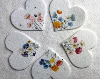 Peut inclure: Cinq ornements en papier en forme de cœur blanc avec des fleurs pressées. Les fleurs sont dans des tons de rose, bleu, jaune et blanc.