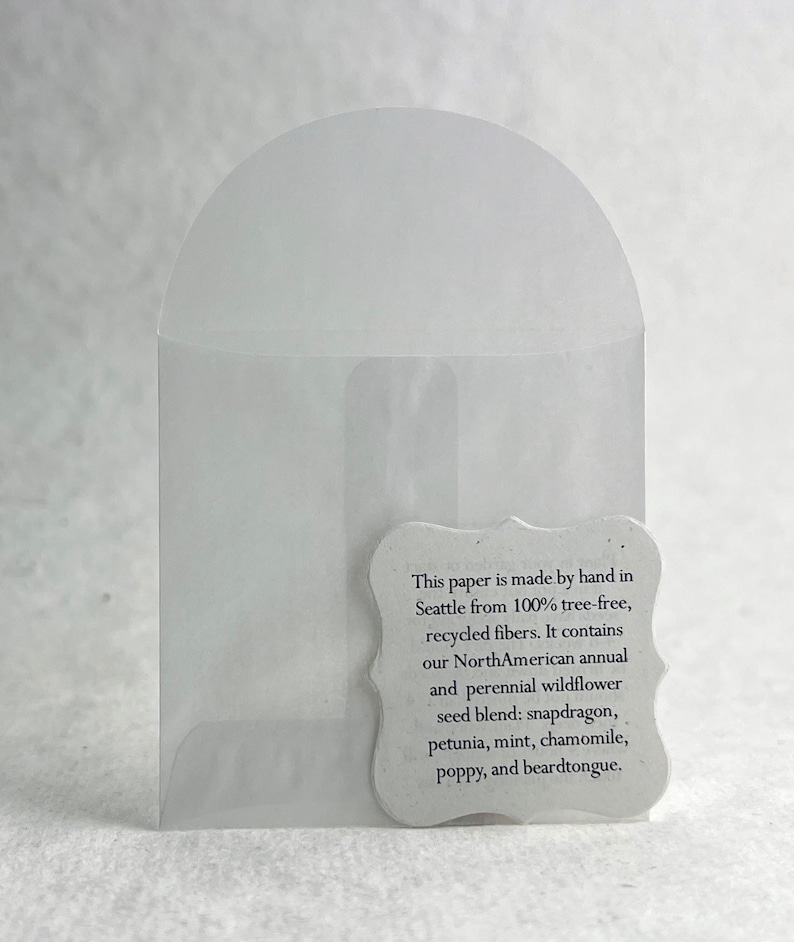 May include: A white paper envelope with a small white tag attached. The tag has text that reads: "This paper is made by hand in Seattle from 100% tree-free, recycled fibers. It contains our NorthAmerican annual and perennial wildflower seed blend: snapdragon, petunia, mint, chamomile, poppy, and beardtongue."