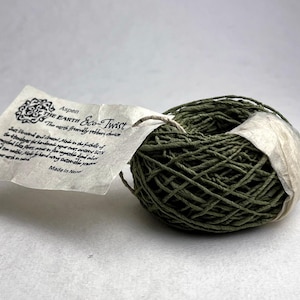 May include: A ball of olive green twisted paper twine wrapped with a white paper band. A rectangular tag is attached, featuring the text "Aspen THE EARTH Eco-Twist" and "The earth friendly ribbon choice," along with additional descriptive text and "Made in Nepal."