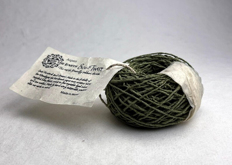 May include: A ball of olive green twisted paper twine wrapped with a white paper band. A rectangular tag is attached, featuring the text "Aspen THE EARTH Eco-Twist" and "The earth friendly ribbon choice," along with additional descriptive text and "Made in Nepal."