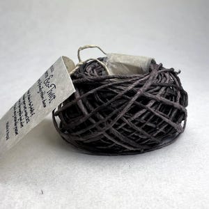 May include: A dark brown, coiled paper twine ribbon labeled Eco-Twist. The textured, twisted paper cord is wound into a compact, rounded bundle. A light-colored, rectangular paper tag is attached to the side, featuring printed text that includes the product name Eco-Twist and origin information.