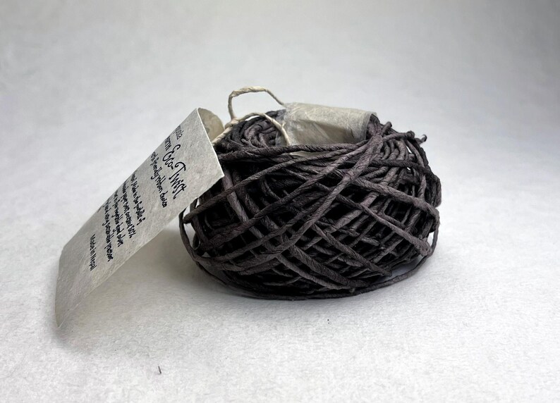 May include: A dark brown, coiled paper twine ribbon labeled Eco-Twist. The textured, twisted paper cord is wound into a compact, rounded bundle. A light-colored, rectangular paper tag is attached to the side, featuring printed text that includes the product name Eco-Twist and origin information.