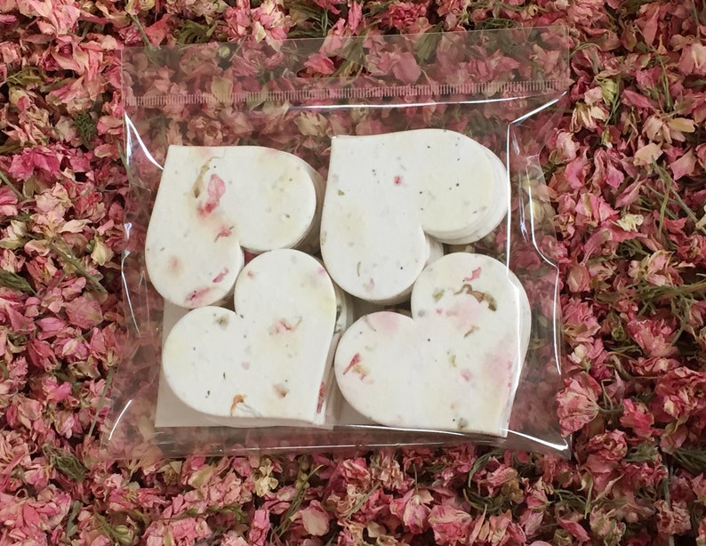 以下が含まれることがあります： 透明なプラスチック製の袋に、白いハート型の紙吹雪が入っています。紙吹雪は乾燥した花びらで作られており、薄いピンク色をしています。袋はピンクの花びらのベッドの上に置かれています。