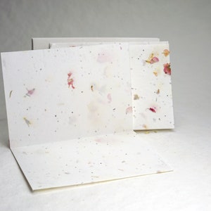 May include: A set of white greeting cards with pink and white flower petals embedded in the paper. The cards are folded in half and have a blank interior.