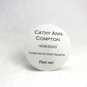 Pu&ograve; includere: Un foglio di carta di semi commemorativo bianco e circolare con testo nero. Il testo recita "CATHY ANN COMPTON 1938-2020 FOREVER IN OUR HEARTS Plant me!" La carta &egrave; leggermente strutturata.