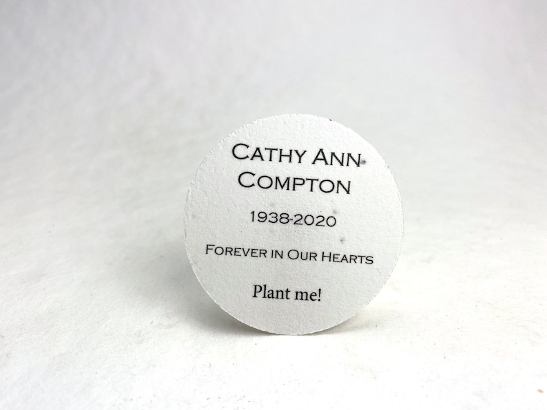Pu&ograve; includere: Un foglio di carta di semi commemorativo bianco e circolare con testo nero. Il testo recita "CATHY ANN COMPTON 1938-2020 FOREVER IN OUR HEARTS Plant me!" La carta &egrave; leggermente strutturata.