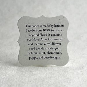 May include: A white paper tag with a scalloped edge and black text. The text reads: "This paper is made by hand in Seattle from 100% tree-free, recycled fibers. It contains our NorthAmerican annual and perennial wildflower seed blend: snapdragon, petunia, mint, chamomile, poppy, and beardtongue."