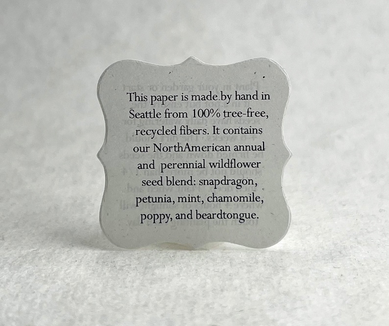 May include: A white paper tag with a scalloped edge and black text. The text reads: "This paper is made by hand in Seattle from 100% tree-free, recycled fibers. It contains our NorthAmerican annual and perennial wildflower seed blend: snapdragon, petunia, mint, chamomile, poppy, and beardtongue."