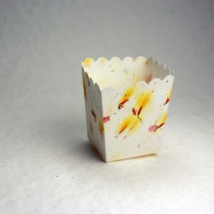 May include: A small, white, scalloped-edge paper box with a decorative pattern of yellow, pink, and red flower petals. The box is empty and sitting on a white surface.