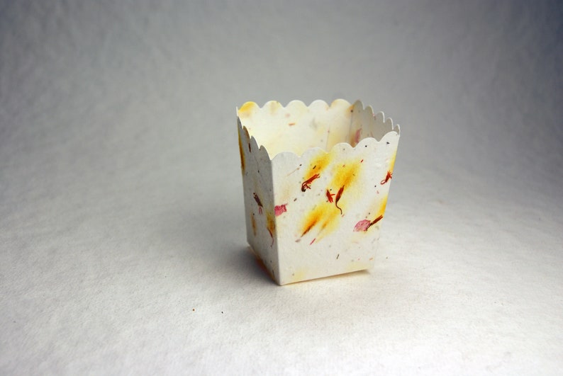 May include: A small, white, scalloped-edge paper box with a decorative pattern of yellow, pink, and red flower petals. The box is empty and sitting on a white surface.