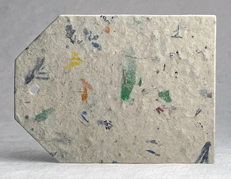 May include: A rectangular tag with a pointed top, made from recycled paper. The tag is off-white with flecks of color, including green, yellow, orange, and blue. A small hole is near the top. The tag is likely used for labeling or crafting.