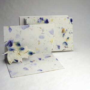May include: A set of three blank cards made of handmade paper with embedded dried flowers. The cards are white with blue and yellow flower petals embedded in the paper. The cards are stacked on top of each other.
