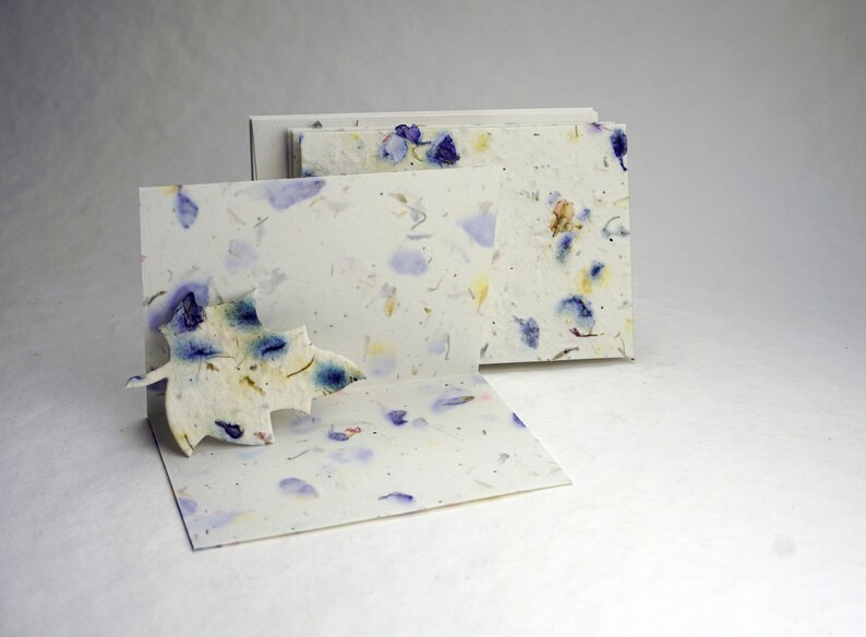 May include: A set of three blank cards made of handmade paper with embedded dried flowers. The cards are white with blue and yellow flower petals embedded in the paper. The cards are stacked on top of each other.