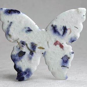 May include: A butterfly-shaped cutout made of white textured paper embedded with small pieces of dried blue and purple flower petals. The paper has a rough, fibrous surface with visible botanical inclusions scattered across the wings.