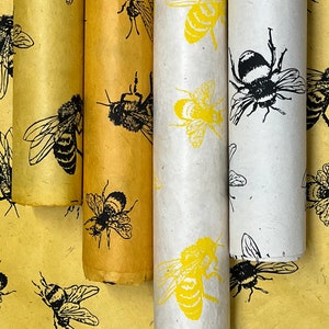 May include: Three rolls of wrapping paper with a bee pattern. The paper is yellow, white, and brown with black bees printed on it. The bees are in different positions and some have yellow accents.