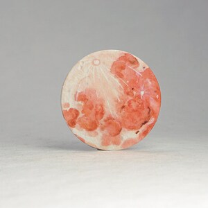 Pu&ograve; includere: Un sottobicchiere rotondo, in stile acquerello, raffigurante la luna. L'opera d'arte presenta sfumature di rosso, arancione e bianco, con crateri dettagliati e un aspetto strutturato. Il sottobicchiere &egrave; un oggetto decorativo.