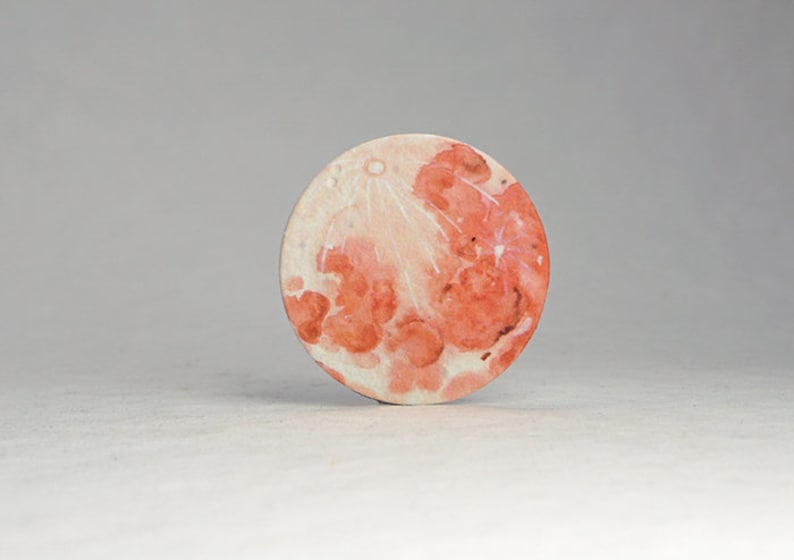 Pu&ograve; includere: Un sottobicchiere rotondo, in stile acquerello, raffigurante la luna. L'opera d'arte presenta sfumature di rosso, arancione e bianco, con crateri dettagliati e un aspetto strutturato. Il sottobicchiere &egrave; un oggetto decorativo.