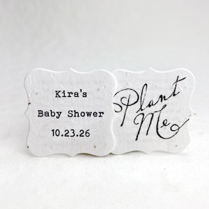 May include: Two white, scalloped-edge paper tags with visible fiber texture. The left tag features black printed text reading Kira's Baby Shower 10.23.26. The right tag features black handwritten-style script text reading Plant Me.