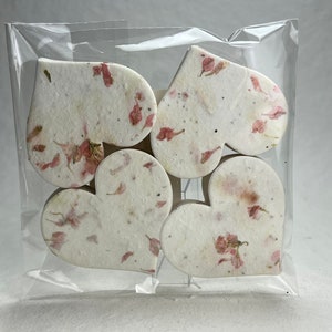 以下が含まれることがあります： ピンクの花びらが埋め込まれた白い紙のハートが6つ入ったパッケージ。ハートは透明なビニール袋に入っています。
