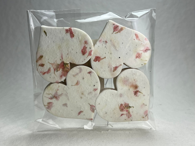 以下が含まれることがあります： ピンクの花びらが埋め込まれた白い紙のハートが6つ入ったパッケージ。ハートは透明なビニール袋に入っています。