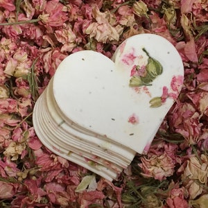 以下が含まれることがあります： 乾燥したピンクの花と緑の葉が埋め込まれた白いハート型の紙吹雪の束
