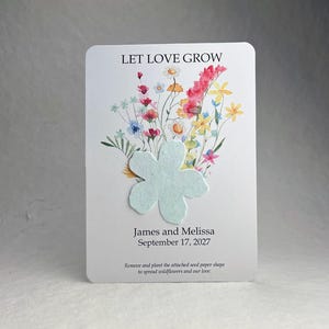 May include: A white rectangular card featuring a watercolor illustration of colorful wildflowers and the text LET LOVE GROW. A light blue flower-shaped seed paper is attached to the center. Below are the names James and Melissa, the date September 17, 2027, and instructions to remove and plant the seed paper.