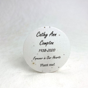Pu&ograve; includere: Un memoriale rotondo di carta di semi bianca con testo nero. Il testo recita "Cathy Ann Compton 1938-2020 Forever in Our Hearts Plant me!". La carta ha piccole macchie. Lo sfondo &egrave; una superficie bianca liscia.