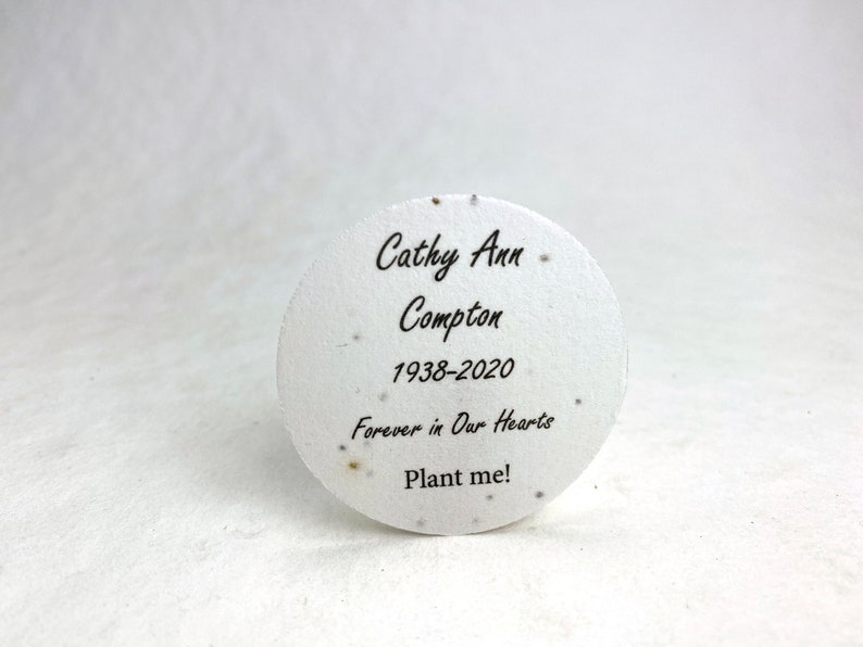 Pu&ograve; includere: Un memoriale rotondo di carta di semi bianca con testo nero. Il testo recita "Cathy Ann Compton 1938-2020 Forever in Our Hearts Plant me!". La carta ha piccole macchie. Lo sfondo &egrave; una superficie bianca liscia.
