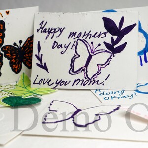 May include: A collection of handmade greeting cards featuring butterfly designs and handwritten messages. The cards are made from recycled paper and have a speckled texture. The messages include "Stay Warm", "Happy Mothers Day! Love you mom!" and "doing okay!"