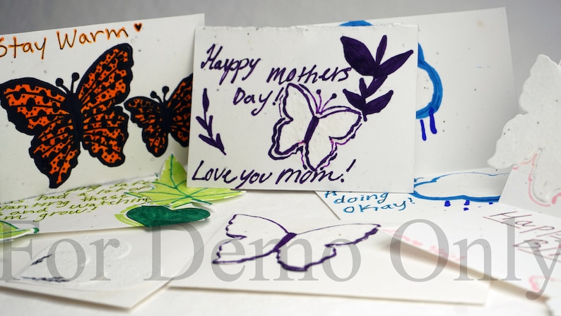 May include: A collection of handmade greeting cards featuring butterfly designs and handwritten messages. The cards are made from recycled paper and have a speckled texture. The messages include "Stay Warm", "Happy Mothers Day! Love you mom!" and "doing okay!"