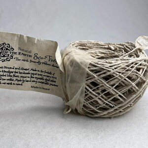 May include: A ball of light beige twine made from recycled lokta fiber, wrapped with a paper tag labeled Natural of the Earth Eco-Twine. The tag includes text detailing its handmade origin in the foothills of the Himalayas and Nepal.