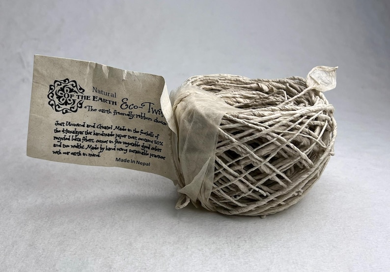 May include: A ball of light beige twine made from recycled lokta fiber, wrapped with a paper tag labeled Natural of the Earth Eco-Twine. The tag includes text detailing its handmade origin in the foothills of the Himalayas and Nepal.