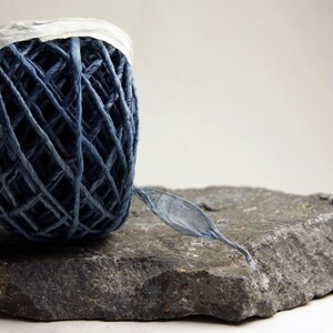 May include: A ball of dark blue raffia ribbon with a crisscross pattern and a white paper band at the top. The ribbon is resting on a flat, textured gray stone, with a single strand of the blue material extending outward from the main ball.