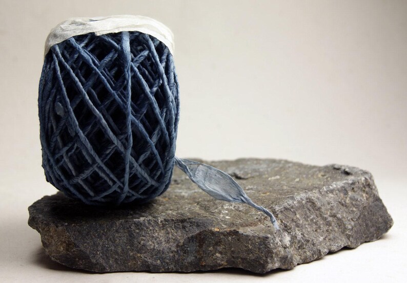 May include: A ball of dark blue raffia ribbon with a crisscross pattern and a white paper band at the top. The ribbon is resting on a flat, textured gray stone, with a single strand of the blue material extending outward from the main ball.