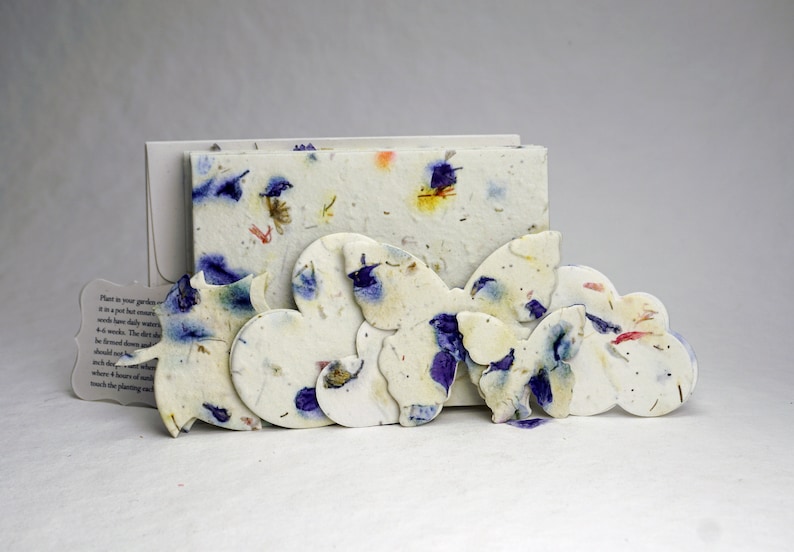 May include: A set of paper butterfly seed paper cards with a white background and blue and purple flower petals. The cards are arranged in a row, with the largest card in the back and the smallest card in the front. The cards are made of recycled paper and are biodegradable.