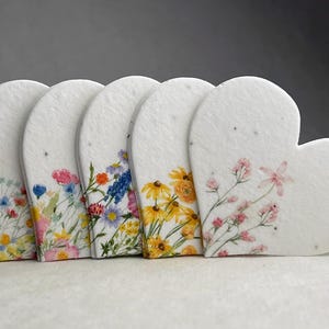 May include: A collection of heart-shaped seed paper cards. Each card features a different watercolor floral design, including yellow, pink, blue, and orange flowers. The cards are arranged in a row, with the rightmost card fully heart-shaped.