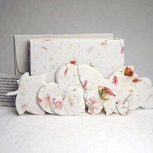 May include: A set of handmade paper cards with a white background and pink flower accents. The cards are shaped like hearts, butterflies, and clouds. The paper is made from 100% recycled fibers.