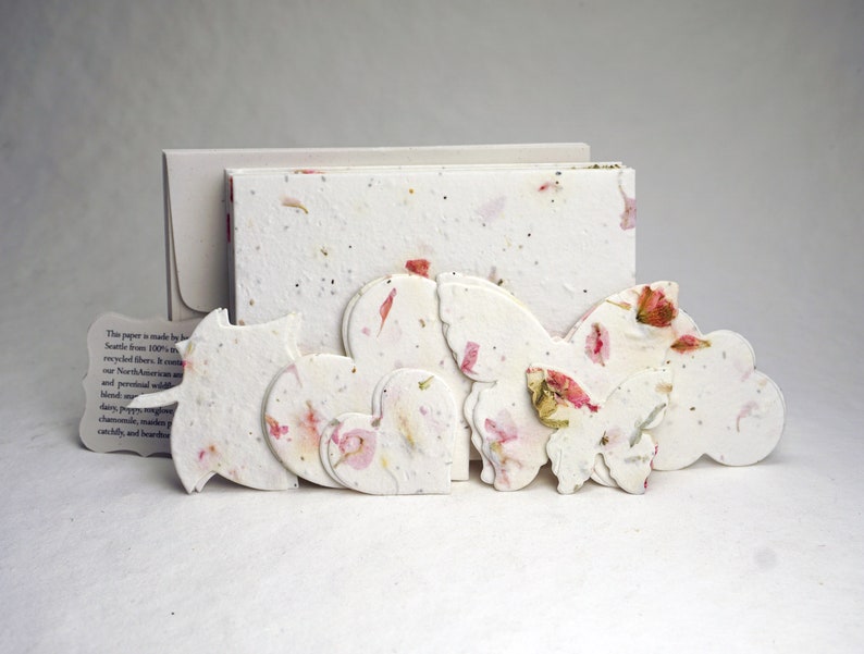 May include: A set of handmade paper cards with a white background and pink flower accents. The cards are shaped like hearts, butterflies, and clouds. The paper is made from 100% recycled fibers.