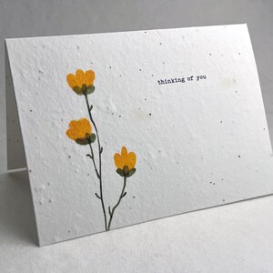 May include: A folded white card with a textured surface. The card features a watercolor illustration of three yellow flowers with green stems. The phrase "thinking of you" is printed in black on the card.