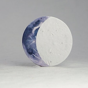 Pu&ograve; includere: Un oggetto decorativo rotondo con un design a mezzaluna. La superficie della luna &egrave; divisa, con una met&agrave; che presenta un lavaggio ad acquerello nei toni del blu e del viola, e l'altra met&agrave; &egrave; bianca e strutturata. L'oggetto &egrave; su uno sfondo neutro.
