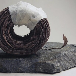 May include: A circular coil of dark brown paper twine secured with a strip of translucent white tape. The twine has a textured, crinkled appearance and a single loose end extending outward from the main roll, resting on a flat, dark gray stone.