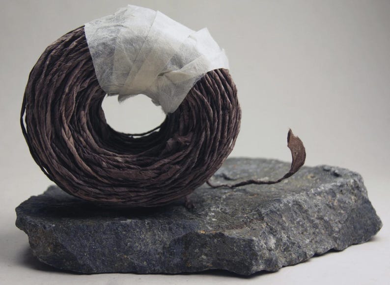May include: A circular coil of dark brown paper twine secured with a strip of translucent white tape. The twine has a textured, crinkled appearance and a single loose end extending outward from the main roll, resting on a flat, dark gray stone.
