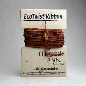 May include: A bundle of brown EcoTwist Ribbon wrapped around a cream-colored paper card. The card features the text EcoTwist Ribbon, 1" Hemlock, 8 Yds., Made in Nepal, 100% Compostable, and additional small print regarding recycled materials and dyes.