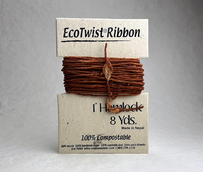 May include: A bundle of brown EcoTwist Ribbon wrapped around a cream-colored paper card. The card features the text EcoTwist Ribbon, 1" Hemlock, 8 Yds., Made in Nepal, 100% Compostable, and additional small print regarding recycled materials and dyes.