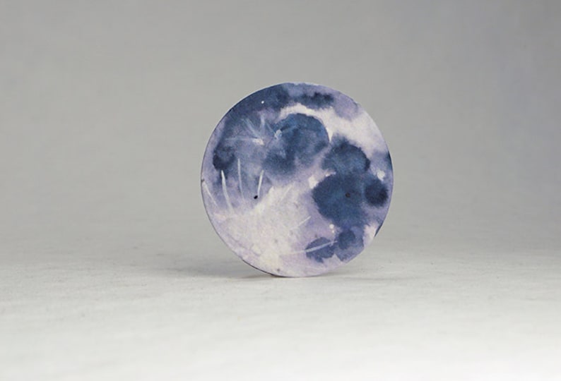 Pu&ograve; includere: Un dipinto rotondo ad acquerello della luna. La luna &egrave; raffigurata in tonalit&agrave; di blu e viola, con riflessi bianchi. L'opera d'arte &egrave; su uno sfondo bianco.