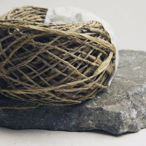 May include: A ball of tan, fibrous twine wrapped in a loose, crisscross pattern, resting on a flat, jagged piece of dark gray stone. A single, pointed, dried leaf-like husk extends from the base of the twine ball to the left side.