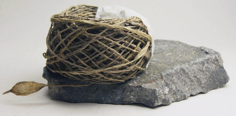 May include: A ball of tan, fibrous twine wrapped in a loose, crisscross pattern, resting on a flat, jagged piece of dark gray stone. A single, pointed, dried leaf-like husk extends from the base of the twine ball to the left side.