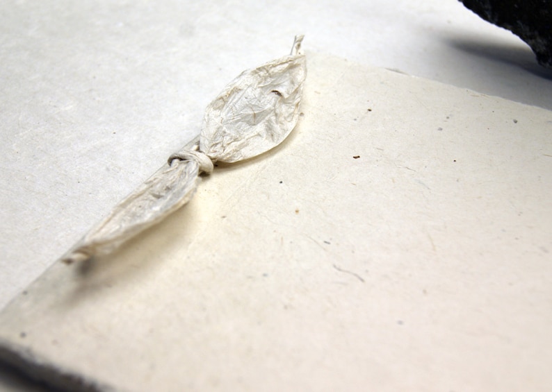 May include: A rectangular, off-white handmade paper card with a textured, fibrous surface. A thin, crinkled, off-white paper strip is tied in a knot around the top edge of the card, creating a decorative accent on the plain, light-colored surface.