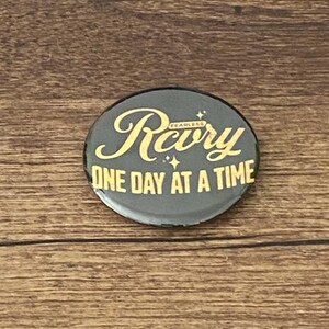 May include: A round, black button with gold lettering. The button reads "Rcvry" above "ONE DAY AT A TIME". The word "FEARLESS" is written in small letters above the "y" in "Rcvry".